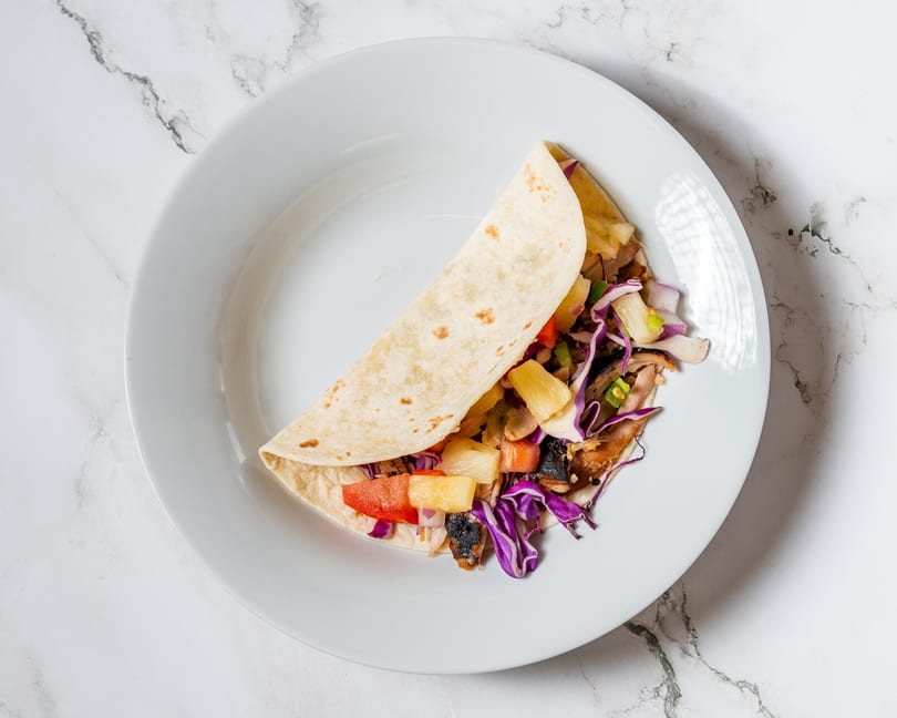 A single taco filled with pineapple, red cabbage, and other vegetables on a white plate.