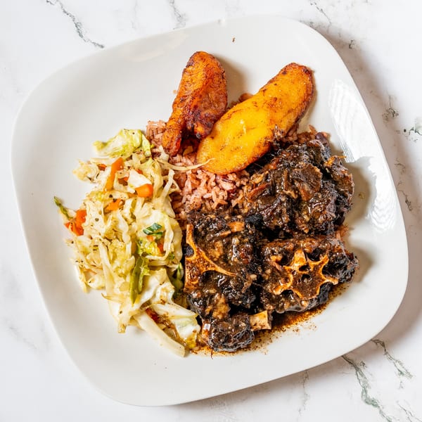 Oxtail stew with rice and peas, fried plantains, and steamed cabbage on a white plate.