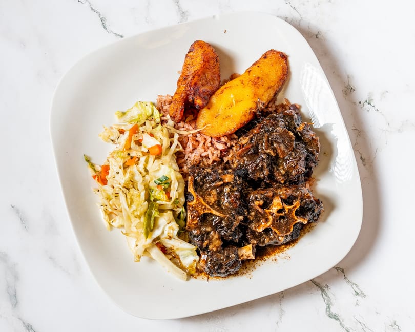 Oxtail stew with rice and peas, fried plantains, and steamed cabbage on a white plate.