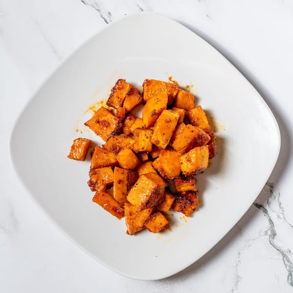 A white square plate filled with glistening, diced sweet potatoes, seasoned and roasted to a golden-brown.