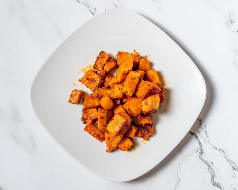 A white square plate filled with glistening, diced sweet potatoes, seasoned and roasted to a golden-brown.