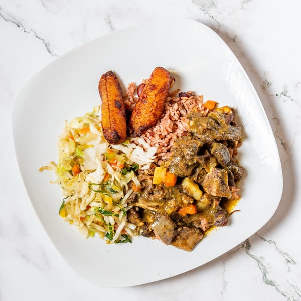 Curried goat with rice and peas, fried plantains, and steamed cabbage on a white plate.