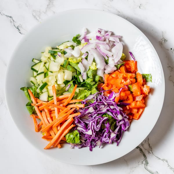 A white bowl filled with colorful salad ingredients: lettuce, cucumbers, red onions, tomatoes, carrots, and red cabbage.