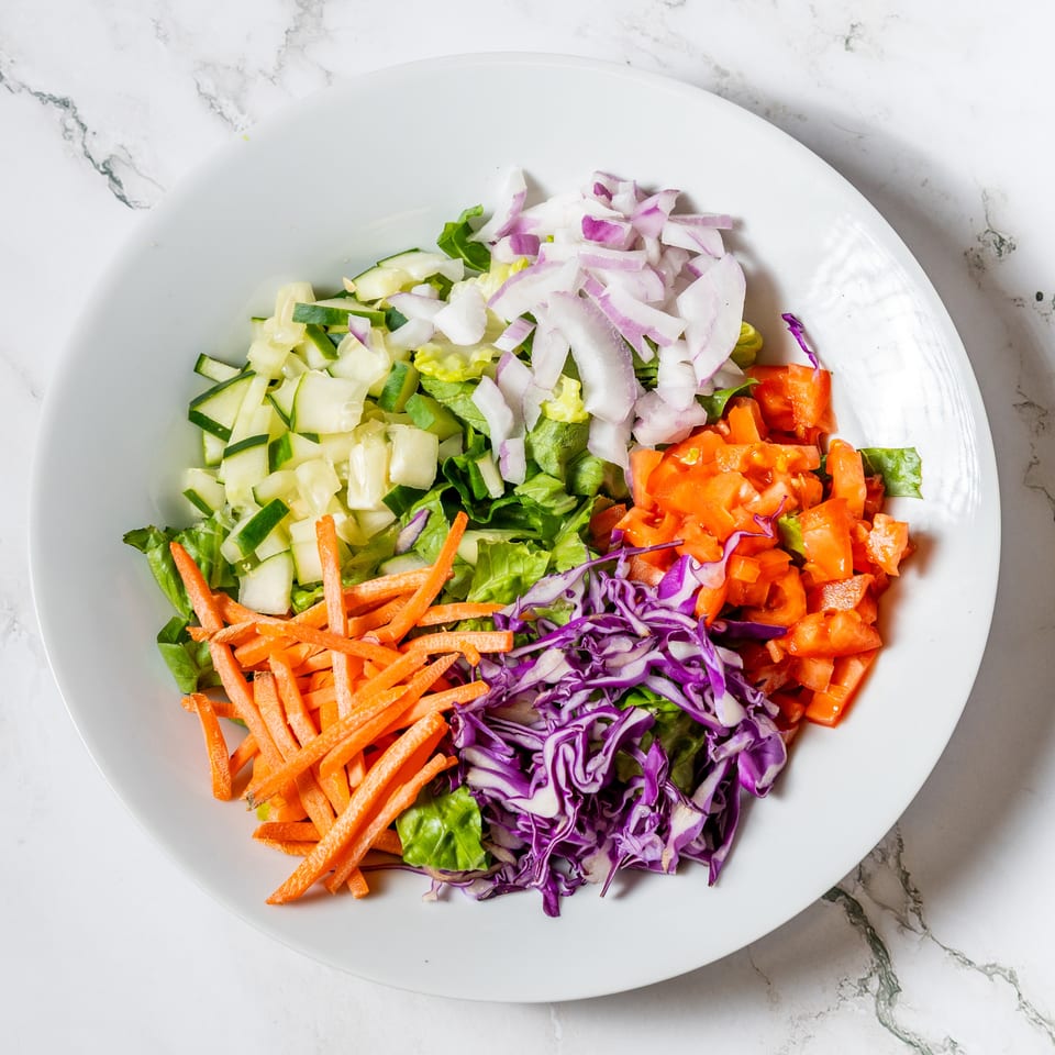 A white bowl filled with colorful salad ingredients: lettuce, cucumbers, red onions, tomatoes, carrots, and red cabbage.