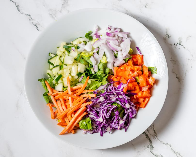 A white bowl filled with colorful salad ingredients: lettuce, cucumbers, red onions, tomatoes, carrots, and red cabbage.