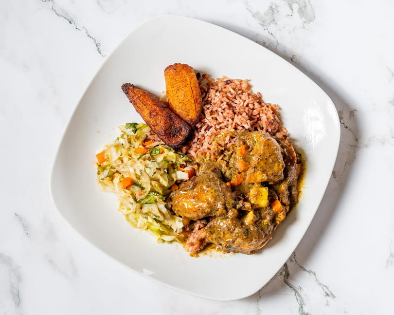 Curry chicken with rice and peas, fried plantains, and steamed cabbage on a white plate.