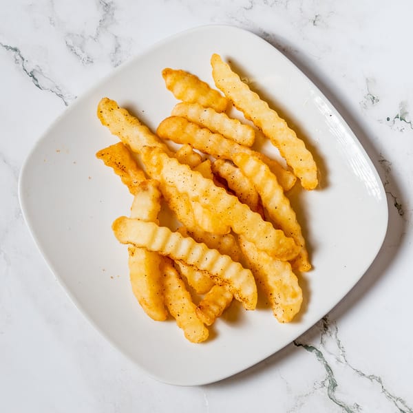 A white plate filled with golden, crinkle-cut french fries, seasoned with pepper, on a marble surface.