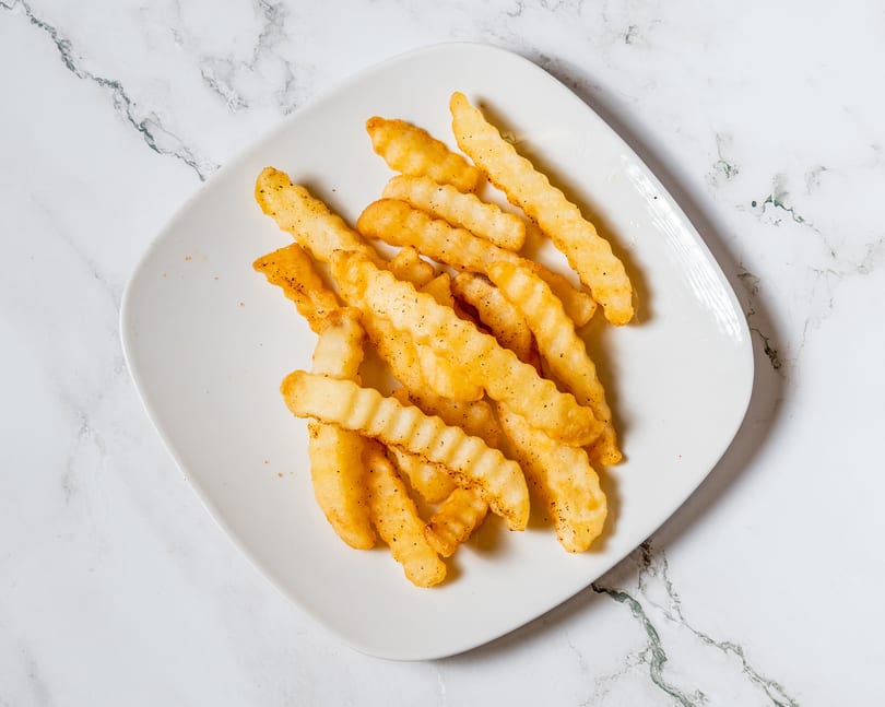 A white plate filled with golden, crinkle-cut french fries, seasoned with pepper, on a marble surface.