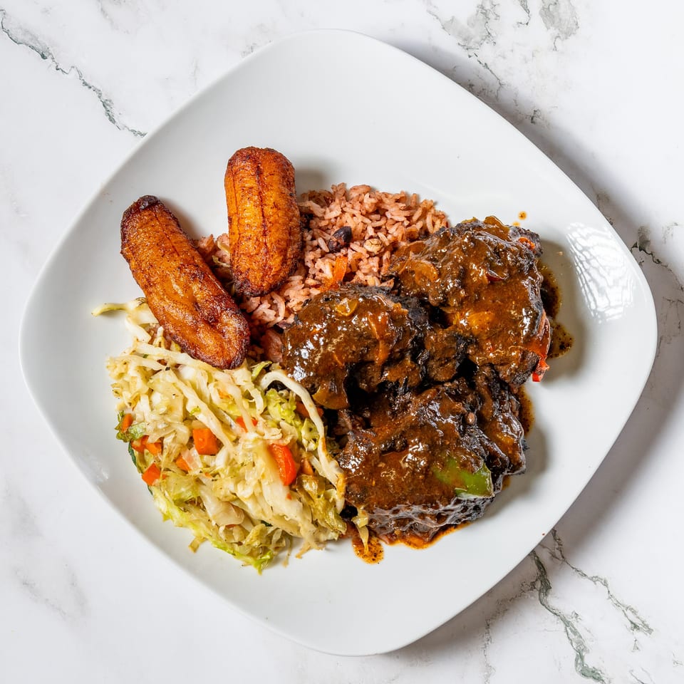 Oxtail stew with rice and peas, fried plantains, and steamed cabbage on a white plate.