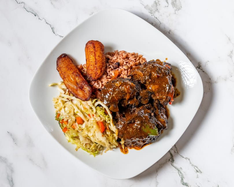 Oxtail stew with rice and peas, fried plantains, and steamed cabbage on a white plate.