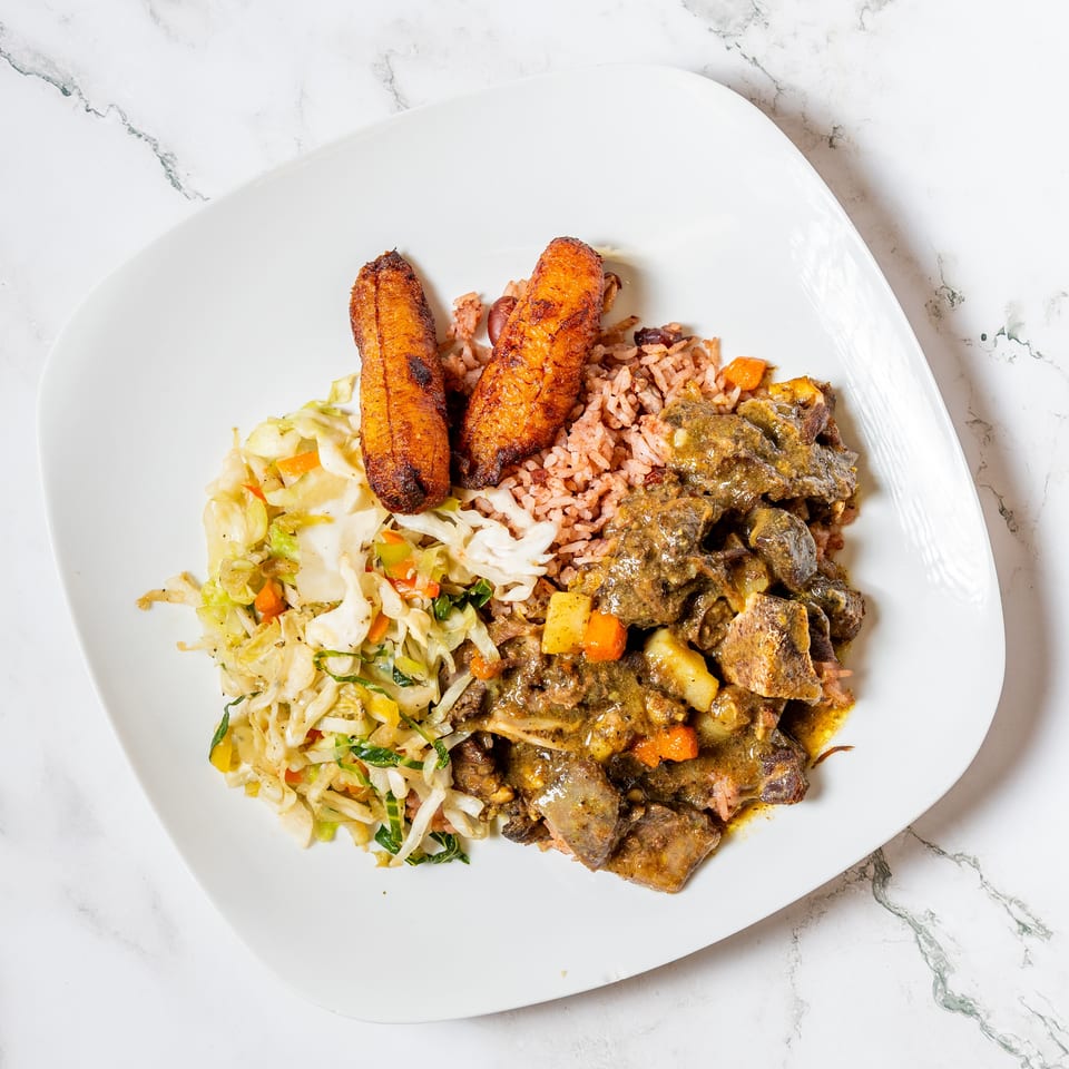 A white plate with Jamaican curry goat, rice and peas, fried plantains, and steamed cabbage.