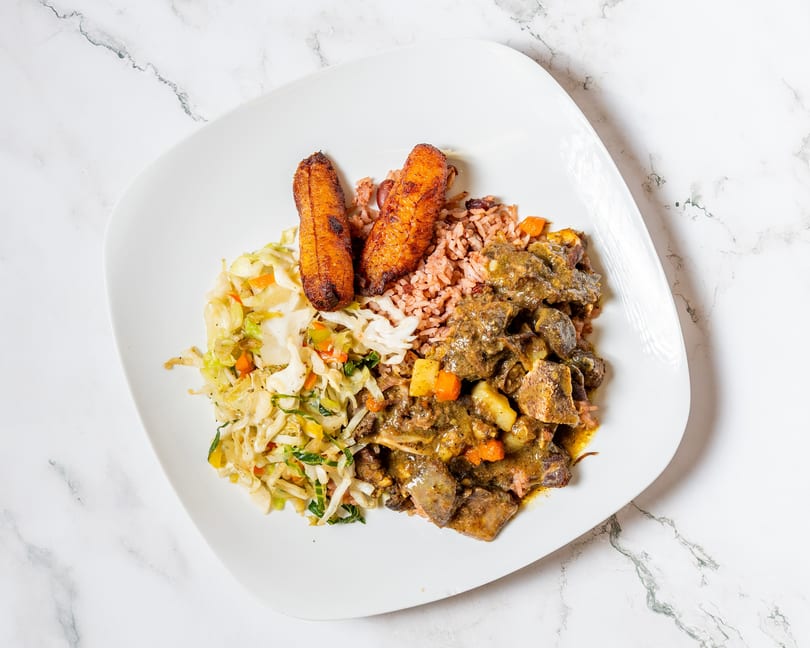 A white plate with Jamaican curry goat, rice and peas, fried plantains, and steamed cabbage.